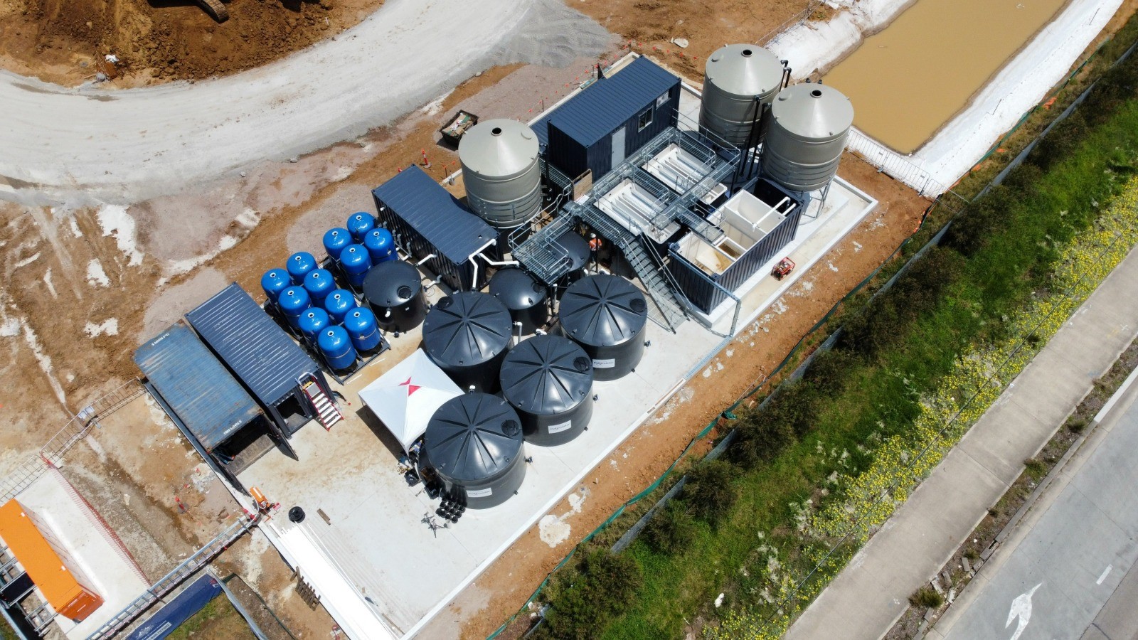 Aerial view of the containerised water treatment plant at Claremont Meadows — clarifier tanks, blue filter vessels and holding tanks laid out beside the settlement pond (photo courtesy of Aquatic Engineering)